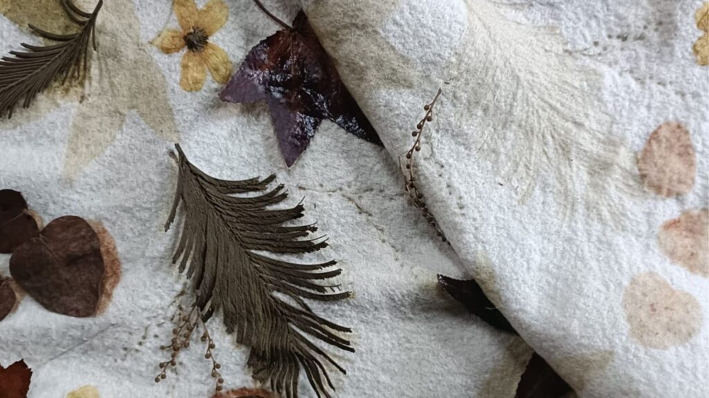 Tela blanca con estampados botánicos en tonos suaves, junto a hojas secas y pequeñas flores prensadas.